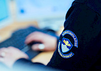 A person typing on a laptop wearing a black jacket with a blue and gold badge on the sleeve.