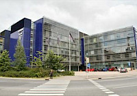 Photo of a modern blue and gray office building with a crosswalk in front. The building has large windows and is multi-storey and rectangular in shape. The building is located on a street corner with a crosswalk in front. There are trees and bushes on the left side of the building. There are cars parked on the street in front of the building. The sky is cloudy and overcast.