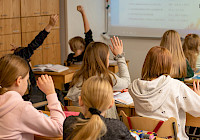 A classroom where students raise their hands. Students sit on desks and chairs made of wood. In the background is a blackboard with text and a wooden wall.