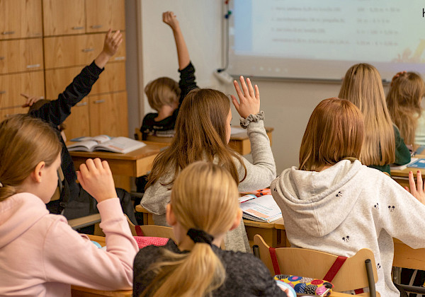 Ett klassrum där eleverna räcker upp handen. Eleverna sitter på skrivbord och stolar av trä. I bakgrunden finns en svart tavla med text och en trävägg.