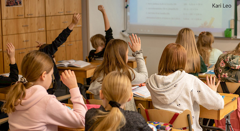 Luokkahuone, jossa oppilaat nostavat kätensä. Oppilaat istuvat puusta valmistetuilla pulpeteilla ja tuoleilla. Taustalla on liitutaulu, jossa on tekstiä, ja puinen seinä.