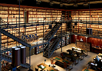 A library with bookshelves, tables and a spiral staircase. The bookshelves are made of wood and full of books. The tables are made of wood and have lamps. The spiral staircase is made of metal and leads to the second floor. The library has a high ceiling with hanging lamps. The floor is made of wood.