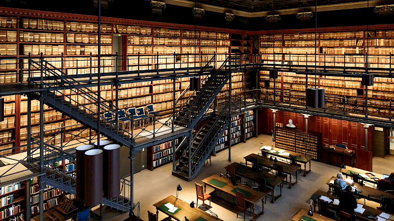 A library with bookshelves, tables and a spiral staircase. The bookshelves are made of wood and full of books. The tables are made of wood and have lamps. The spiral staircase is made of metal and leads to the second floor. The library has a high ceiling with hanging lamps. The floor is made of wood.