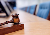 A wooden mallet resting on a wooden table in a courtroom. The mace is in the foreground and in focus. The background is out of focus and consists of a row of blue chairs and a window. The table is polished and you can see the reflection of the mallet on it.