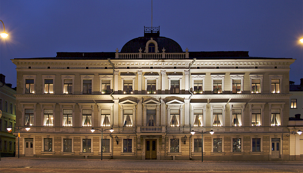 Bilden. På bilden finns en byggnad som används av Högsta domstolen i Helsingfors. En stor vit trevåningsbyggnad med många fönster. Huset och fönstren är upplysta. Framför bilden syns kullerstensgatan och himlen är mörkblå.