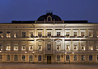 Illustration. The picture shows a building used by the Supreme Court in Helsinki. A large light three-story building with lots of windows. The house and windows are illuminated. In front of the picture you can see a cobblestone street and the sky is dark blue.