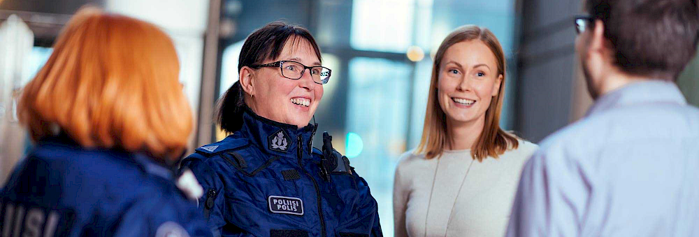 Ryhmä ihmisiä kirkkaassa toimistoympäristössä. Ryhmään kuuluu kaksi poliisia ja kaksi siviiliä. Poliisit käyttävät tummansinisiä univormuja, joissa on selässä teksti ‘POLIISI’. Siviilit ovat pukeutuneet rentoihin vaatteisiin. Ihmisten kasvot on sumennettu yksityisyyden vuoksi. Taustalla on suuri ikkuna ja pöytä.