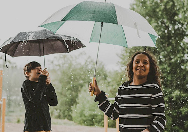 Två personer som går i regnet med gröna och svarta paraplyer. Människor går på en stig med träd i bakgrunden.