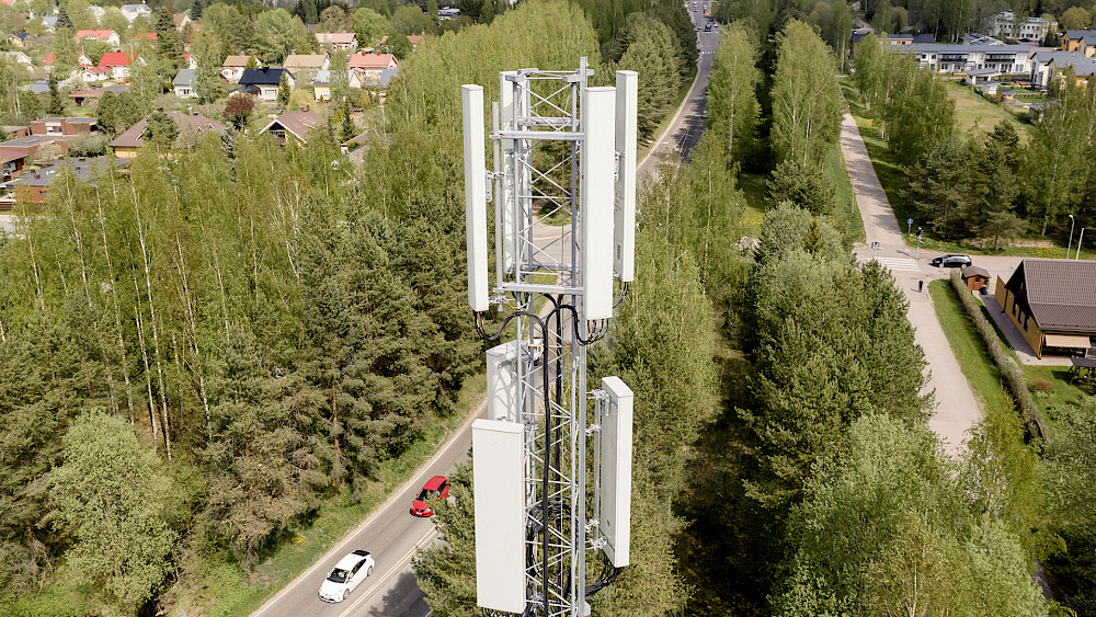 Flygfoto över ett mobiltelefontorn i ett skogsområde, med en väg och hus i bakgrunden. Tornet är högt och vitt med flera antenner. Tornet ligger intill vägen där den röda bilen kör. Det finns hus och andra byggnader i bakgrunden. Träden är gröna och himlen är blå.