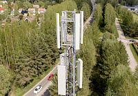 Aerial view of a cell phone tower in a wooded area, with a road and houses in the background. The tower is tall and white with multiple antennas. The tower is located next to the road where the red car drives. There are houses and other buildings in the background. The trees are green and the sky is blue.