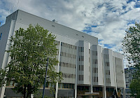 A photograph of a white building with trees in front and a blue sky in the background. The building is modern, minimalist and has a flat roof and rectangular windows. There are trees and bushes around the building. The sky is blue and there are a few clouds. In the foreground are two out of focus people walking.