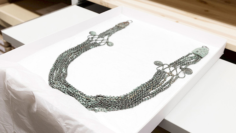 Valokuva kaulakorusta, joka on tehty pienistä metallirenkaista ja kolikoista valkoisella pinnalla. Kaulakoru on muodoltaan U-kirjaimen muotoinen ja siinä on lukko yläosassa.