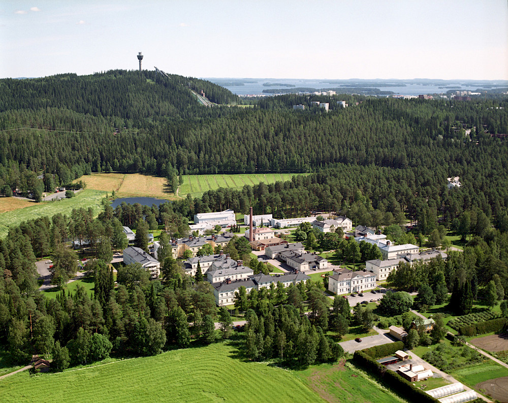 Ilmakuva kaupungista, jossa on vihreitä peltoja ja järvi taustalla. Kaupunki on ympäröity puilla ja vihreydellä. Kaupungissa on sekoitus moderneja ja perinteisiä rakennuksia. Kaupungissa on korkea torni taustalla. Järvi on näkyvissä taustalla.