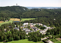 Aerial view of a city with green fields and a lake in the background. The city is surrounded by trees and greenery. The city has a mix of modern and traditional buildings. The city has a tall tower in the background. The lake is visible in the background.