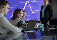 Illustration. Several people working at the desk in the meeting room. The woman stands behind the table. A graphical statistical curve is projected on the wall.