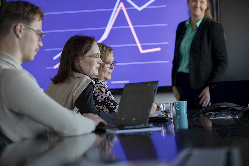 Kuvituskuva. Useita henkilöitä työskentelemässä työpöydän ääressä kokoustilassa. Nainen seisoo pöydän takana. Seinälle on heijastettu graafinen tilastokäyrä.