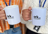 Two hands holding white mugs with 'ork' written on them. The mugs have a blue logo with white letters. The background is out of focus and consists of a person wearing an orange shirt and blue jeans. Person's hands are focused and holding mugs.