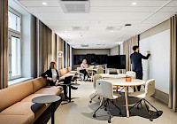 A modern office space where a man is writing on a whiteboard and two women are sitting on a sofa. The office space is well lit with large windows and light walls. The furniture is modern and minimalist, including a beige sofa, white tables and black chairs. The man is dressed in a black suit and writes on the blackboard with a marker. The women are sitting on a sofa and seem to be having a conversation.