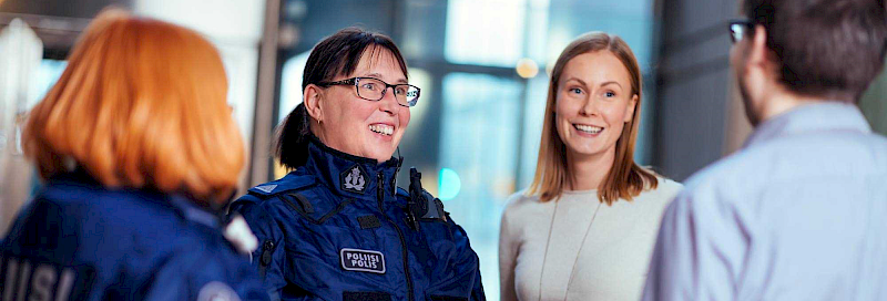 Ryhmä ihmisiä kirkkaassa toimistoympäristössä. Ryhmään kuuluu kaksi poliisia ja kaksi siviiliä. Poliisit käyttävät tummansinisiä univormuja, joissa on selässä teksti ‘POLIISI’. Siviilit ovat pukeutuneet rentoihin vaatteisiin. Ihmisten kasvot on sumennettu yksityisyyden vuoksi