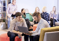 Two people working on a laptop in a modern office space, blurred people in the background. People sit on red and orange stools. The laptop is open and appears to be in use. The background consists of blurred people and bookshelves. The color scheme is mainly white and blue.