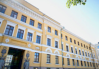 Photo of a yellow building with white decorations and a blue sky in the background. The building has three floors and a basement. The building has a flat roof and a toothed molding. The windows are rectangular and have white frames. The entrance is in the middle of the building.