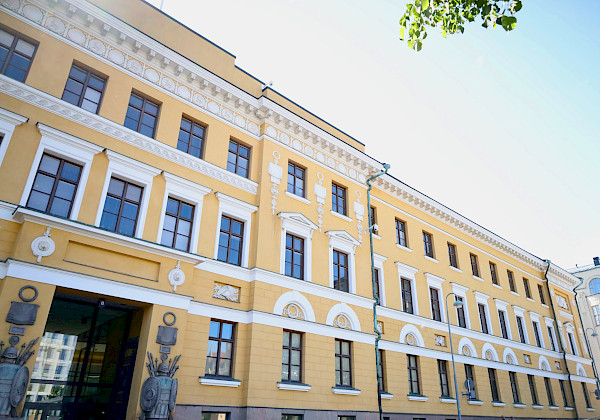 Valokuva keltaisesta rakennuksesta, jossa on valkoisia koristeita ja taustalla sininen taivas. Rakennuksessa on kolme kerrosta ja kellarikerros. Rakennuksella on tasakatto ja hammaslista. Ikkunat ovat suorakulmaisia ja niissä on valkoiset kehykset. Sisäänkäynti on rakennuksen keskellä.