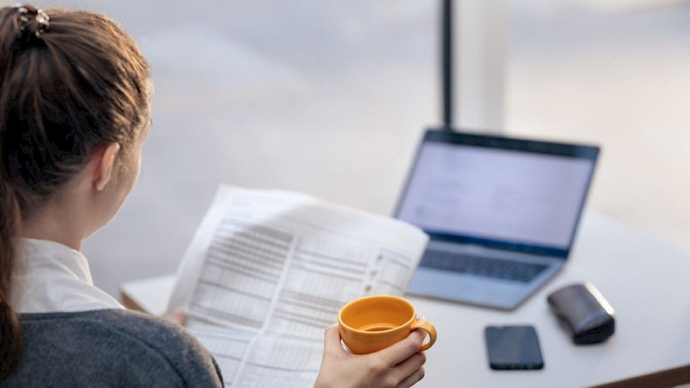 En person arbetar på en bärbar dator medan han håller en orange kaffekopp. En person sitter vid ett bord med en telefon och papper utspridda. Personen är klädd i en grå tröja och håret är bundet i en bulle. I bakgrunden finns ett fönster som visar himlen och molnen.
