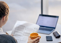 A person is working on a laptop while holding an orange coffee cup. A person is sitting at a table with a phone and papers scattered around. The person is wearing a gray sweater and her hair is tied in a bun. In the background is a window showing the sky and clouds.