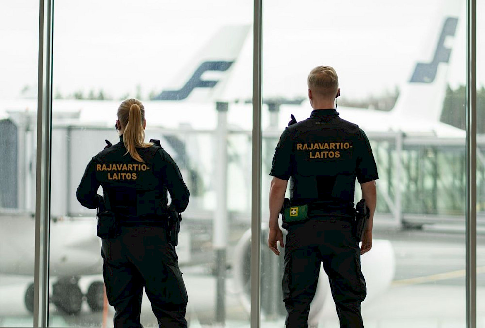 Kaksi rajavartijaa seisoo lentokentän ikkunan edessä. Virkailijat käyttävät mustia univormuja.Virkailijat seisovat selkä kameraan päin. Ikkunasta näkyy lentokone ja lentokentän kiitorata.