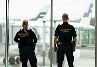 Two border guards are standing in front of the airport window. The clerks wear black uniforms. The clerks stand with their backs to the camera. An airplane and the airport runway can be seen from the window.