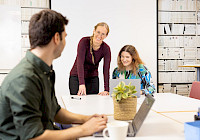 Three people in a meeting in the office, sitting at a table with a laptop, a coffee mug and a potted plant. In the background is a blackboard and a bookshelf.