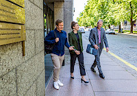 Tre personer går på trottoaren bredvid en byggnad. Byggnaden är modern och gjord av betong. Människor är klädda i business casual stil. Det finns träd och en gata i bakgrunden.