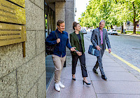 Three people are walking on the sidewalk next to a building. The building is modern and made of concrete. People are dressed in business casual style. There are trees and a street in the background.