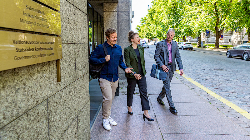 Kolme ihmistä kävelee jalkakäytävällä rakennuksen vieressä. Rakennus on moderni ja betonista. Ihmiset ovat pukeutuneet business casual -tyyliin. Taustalla on puita ja katu.