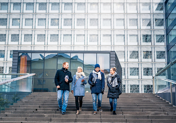 Bilden. En grupp personer går nerför yttertrappan och pratar med varandra. I bakgrunden ser man en stor vit byggnad med många fönster.