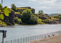 The picture shows a rocky coast with a historic fortress in the background. The fortress is made of stone and grass grows on it. The fortress is located on a rock. The water is deep blue and calm. The sky is blue and there are a few clouds. In the foreground is a metal fence. There is a tree branch in the upper left corner of the picture.