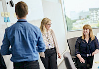 Three people in a modern office space with a window and post-it notes on the wall. People are dressed in business casual style. The person on the left is standing and the other two are sitting.