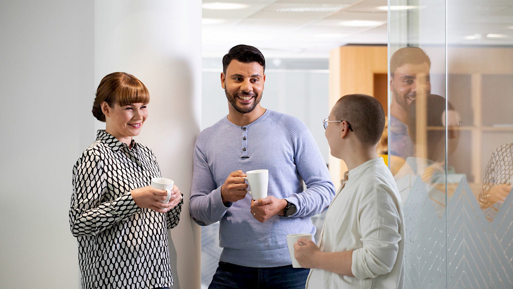 En grupp människor står på ett kontor, håller kaffekoppar och pratar. I bakgrunden finns en kontorsmiljö med skrivbord och fönster. Människor är klädda i business casual stil. Människor håller vita kaffekoppar.