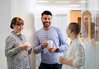 A group of people are standing in an office, holding coffee cups and talking. In the background is an office environment with a desk and a window. People are dressed in business casual style. People are holding white coffee cups.