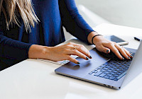 A person is typing on a laptop on a table with a phone and notepads.