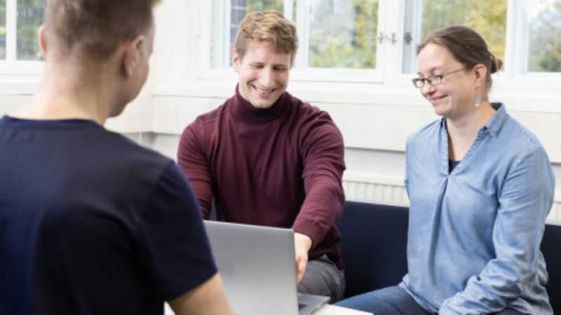 Kolme henkilöä toimistotilassa tietokoneen äärellä. Taustalla valkoinen seinä, jossa ikkunoita, joista näkyy lehtipuita.