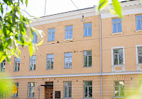 The facade of the yellow building. The building has three floors and plenty of windows. In the foreground you can see the leaves of a deciduous tree.