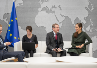 Four people are sitting in a conference room with a world map on the wall and the flags of the European Union and Finland in the background. People are sitting on white chairs and there are two white coffee tables in front of them. One person is holding the phone, another is using a laptop, and the third is holding a notebook.