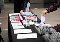 Fair table at the event. There are brochures and pens on the table. The table is covered with a black tablecloth. Brochures come in different colors and sizes. The pens are black and in a holder. There are two people in the background, one of whom is holding a brochure. In the background is a gray carpet and a white wall with a sign.