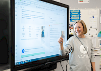 Illustration. A smiling person stands in front of a large flat screen TV and shows graphic patterns and text displayed on it. Papers and posters of different colors are attached to the wall at the back. The person in the picture is wearing a grey shirt with the logo and name of the Valteri school printed on it.