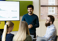 En grupp människor i ett möte med en presentation på skärmen i bakgrunden.