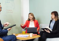 Three people in a meeting room. One is holding a notebook, one is holding a tablet, and one is holding a computer. In the background, a whiteboard with the words Customer in focus. People have coffee cups in front of them.