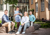 A group of people are sitting on a rock in front of a building. The building is pale yellow with white windows and a green garden in front. People are dressed in casual clothes.