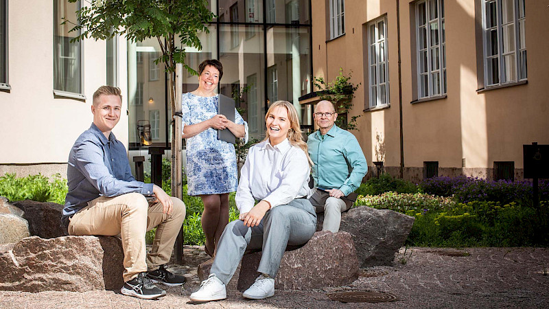 Ryhmä ihmisiä istuu kalliolla rakennuksen edessä. Rakennus on vaaleankeltainen, siinä on valkoiset ikkunat ja sen edessä on vihreä puutarha. Ihmiset ovat pukeutuneet rentoihin vaatteisiin.