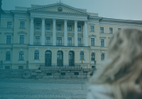 A large neoclassical building. The image is tinted with a blue-green tone. The building has a large pediment with a coat of arms in the center. The building is three stories high. The building is located on a cobblestone street. In the foreground of the picture is the upper body of the person photographed from behind.